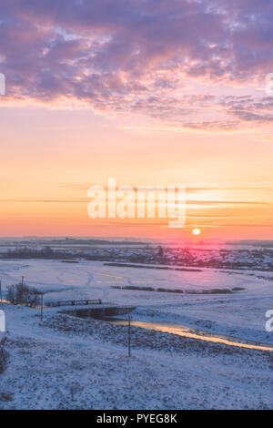 Die malerische Landschaft mit Dawn am frühen Morgen über einen ländlichen Bereich mit einem See und einem hellen roten Sonne am Horizont. Winter Stockfoto