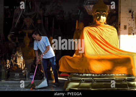 Ein Mitarbeiter der ein Geschäft, in dem buddhistische Devotionalien in Bamrung Muang Rd., Bangkok, Thailand, fegt den Gehweg vor seinem Laden Stockfoto