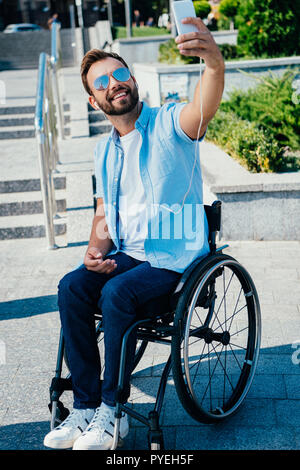 Schöner Mann im Rollstuhl unter selfie mit Smartphone und Anhören von Musik auf der Straße Stockfoto