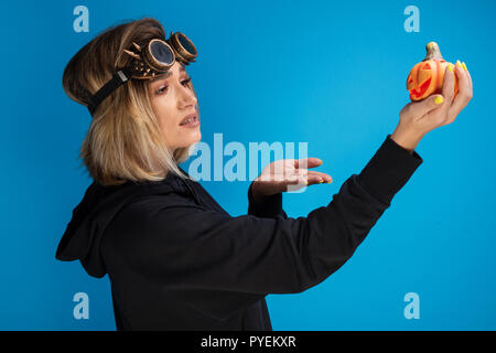 Portrait von Goth Girl tragen Steam Punk Brille wunderte sich ein orange Kürbis geschnitzt. Gothinc posiert im Studio auf blauem Hintergrund Stockfoto