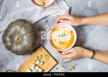 Weibliche Hände halten eine Schüssel Kürbiscremesuppe auf konkreten Hintergrund. Ansicht von oben Stockfoto