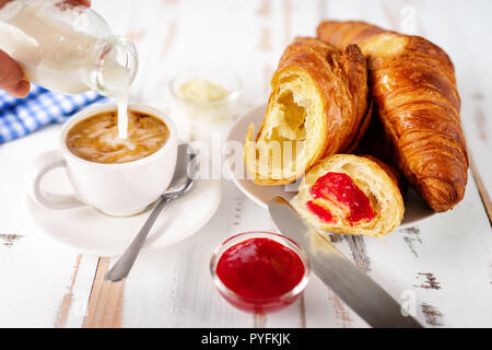 Gießen Milch in die Tasse Kaffee neben einer Platte mit Croissants auf weißen Tisch Stockfoto