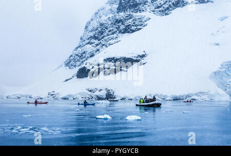 Über das Motorboot mit Touristen und Kajaks in der Bucht mit Felsen und Gletscher im Hintergrund Schneefall, in der Nähe von Almirante Brown, Antarktische Halbinsel Stockfoto