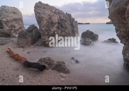 Abenddämmerung am Strand unmittelbar nach Sonnenuntergang, grossen Felsen, türkisfarbenen Wasser des Ionischen Meeres, milchig Wellen und große Stück firebrand auf Kiesel Stockfoto