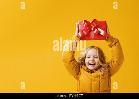 Happy little Girl Vorschüler in einem Winter Jacke hält eine Box mit einem Geschenk, das über den Kopf. Das Konzept von Geben und Empfangen von Geschenken. Gelb auf einem Ihr Stockfoto