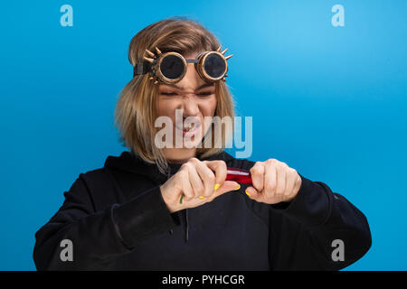 Mädchen mit Steam Punk Brille reißen eine rote Chili, während sie wütend. Portraitfotos im Studio vor blauem Hintergrund Stockfoto