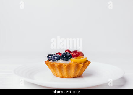 Close-up schöne Törtchen mit Beeren an der Platte. Sauberes Bild elegante Cupcake dekoriert mit Heidelbeeren, Johannisbeeren und Mango auf weißem Hintergrund mit Stockfoto