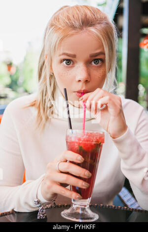 Junge blonde Frau, mit Sommersprossen wundern und Trinken red Cocktail mit Beeren und Minze in Outdoor Restaurant Stockfoto