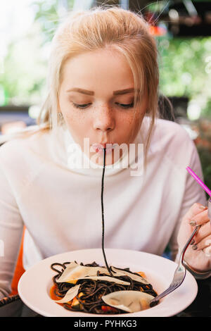 Junge schöne Frau essen schwarze Nudeln mit Meeresfrüchten und Tintenfisch Tinte in das Restaurant im Freien. Lustig und schön. Stockfoto