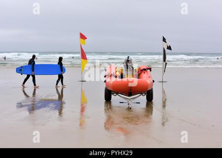 Menschen, die ein blaues Surfbrett hinter RNLI Strand Sicherheit Flaggen und aufblasbare Rettungsboote, Perranporth Beach, Cornwall, England, Großbritannien Stockfoto