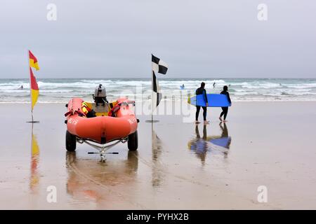 Menschen, die ein blaues Surfbrett hinter RNLI Strand Sicherheit Flaggen und aufblasbare Rettungsboote, Perranporth Beach, Cornwall, England, Großbritannien Stockfoto