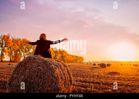Gerne Frau mittleren Alters auf haystack im Herbst Feld und das Gefühl, sich mit den Armen eröffnet. Entspannen und genießen Sie die Natur bei Sonnenuntergang Stockfoto