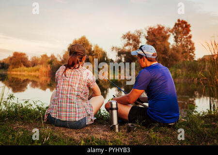Junges Paar angeln und Tee trinken auf dem Fluss bei Sonnenuntergang. Menschen entspannen und Chillen am See sitzen im Freien Stockfoto