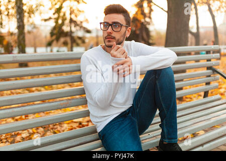 Junge schöne hipster Kerl sitzt im Herbst Park auf einer Bank. Alleine langweilen. Ein Student Spaziergänge im Park. bärtigen Mann in Gläsern. Stockfoto