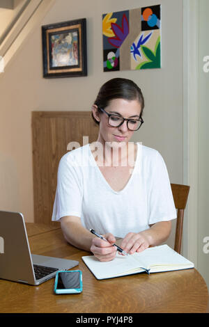 Junge Frau Künstler schreiben im Journal mit Handy und Laptop und Gemälden an der Wand hinter Stockfoto