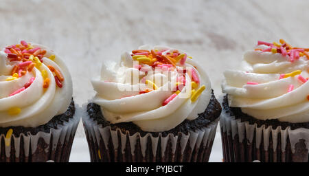 3 chocolate Cupcakes wit Vanille Glasur und Streuseln auf hellem Holz Hintergrund Nahaufnahme Stockfoto