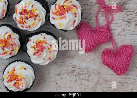 Schoko Cupcakes mit weißem Zuckerguss und besprüht, Gestrickte rosa Herzen auf hellem Holz Hintergrund Stockfoto