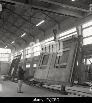 1960, männlichen Arbeitnehmer in Haken mit einer Riemenscheibe zu einem großen Pre-betonfassade Panel mit Fensterrahmen aus Gusseisen für den Gebrauch im Hochhaus oder Bürogebäude, England, UK. Diese Art der 'modernen' Bauweise wurde sehr beliebt in dieser Zeit, wie es erlaubt große Gebäude schnell konstruiert werden und zu geringeren Arbeits- und Materialkosten, da große Teile der Gebäude wie der Fassadenplatten aus, kann sein. Stockfoto