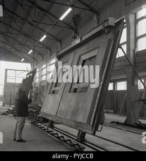 1960, männlichen Arbeitnehmer in Haken mit einer Riemenscheibe zu einem großen Pre-betonfassade Panel mit Fensterrahmen aus Gusseisen für den Gebrauch im Hochhaus oder Bürogebäude, England, UK. Diese Art der 'modernen' Bauweise wurde sehr beliebt in dieser Zeit, wie es erlaubt große Gebäude schnell konstruiert werden und zu geringeren Arbeits- und Materialkosten, da große Teile der Gebäude wie der Fassadenplatten aus, kann sein. Stockfoto