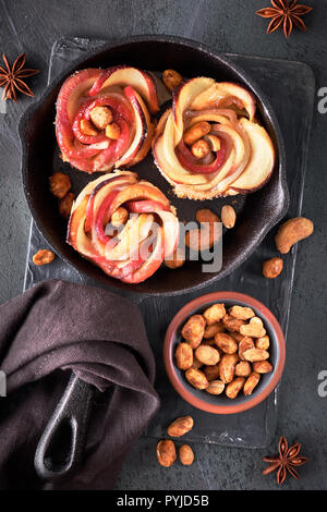 Drei selbstgemachten Blätterteig Gebäck mit geformten rose Apfelspalten gebacken in Gusseisen Skillet. Top lag auf dunklem Hintergrund mit Bettwäsche Handtuch und karamellisierten Mutter Stockfoto