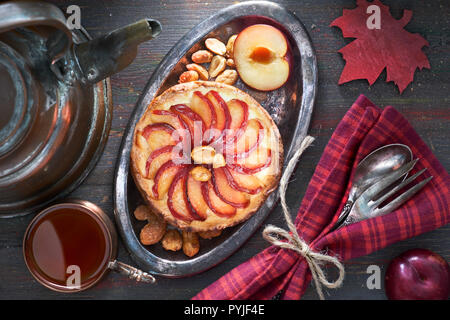 Hausgemachte Pflaumen Tarte mit Hälfte frische Pflaumen- und Erdnüsse auf Metallplatte mit Geschirr, schwarzer Tee und Kaffee Wasserkocher auf dunklen Holztisch, oben legen Stockfoto