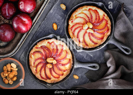 Hausgemachte Torten Streusel mit Pflaume Schichten gebacken in kleine Eisen skillets. Top lag auf dunkel strukturierten Hintergrund mit einigen Pflaumen, Erdnüssen und rote Blätter Stockfoto