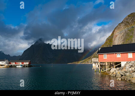 Rainbow ofer rote Häuser Rorbuer in der Reine auf den Lofoten, Norwegen mit roten rorbu Häuser, Wolken, regnerisch blauer Himmel und Sonnenschein. Brücken und Berge in der Rückseite Stockfoto