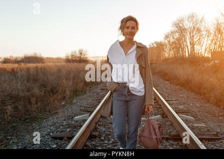 Eine junge Frau in einem weißen Hemd, jeans beige Regenmantel und genießt die Natur, Wandern entlang der Eisenbahngleise rund um blauen Himmel. Das Konzept der livestyle ein Stockfoto