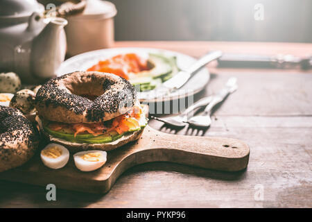 Hausgemachte sandwich Bagel mit Lachs, Avocado, frischen Käse und gekochte Wachteleier auf dunklen Holzmöbeln im Landhausstil Hintergrund. Frühstück oder ein Snack preparat Stockfoto