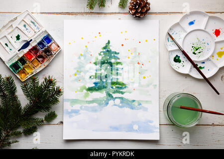 Handgefertigte watercolor Christmas Card auf dem Arbeitstisch mit Wasserfarben, Pinsel, Palette und Tannenzweigen und Tannenzapfen. Ansicht von oben Stockfoto
