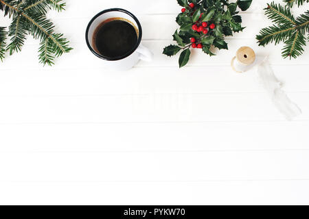 Weihnachten Komposition. Winter Frühstück noch leben Szene. Rahmen der Fir Tree Branches, Holly Blätter und Beeren, Seidenband und Emaille Tasse Kaffee. Weiße Holztisch Hintergrund. Flach, Ansicht von oben, kopieren. Stockfoto