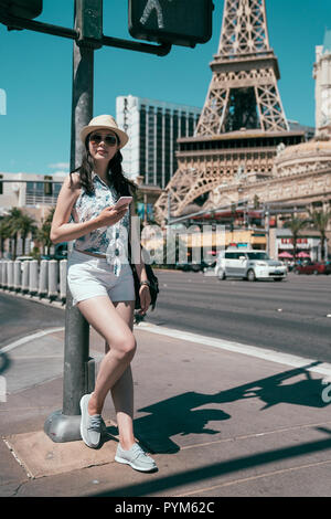 Sorglose Mädchen spielen online Spiel App auf dem Smartphone während der Wartezeit die Straße im Sommer zu überqueren. Tower in Las Vegas im blauen Himmel entfernt. Zuversichtlich w Stockfoto