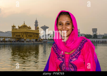 Das Porträt einer lächelnden jungen weiblichen Sikh devotee, in roten Tüchern gekleidet, vor dem Harmandir Sahib, der Goldene Tempel Stockfoto