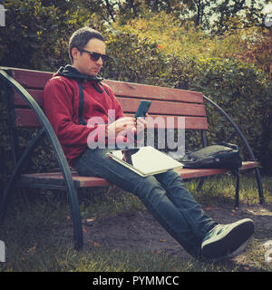 Junge hipster Kerl Kontrolle Benachrichtigung auf Mobiltelefon, während Entfernung Arbeiten am Laptop, im Stadtpark, im Sommer Tag sitzen. schöner Mann chatt Stockfoto