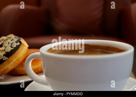 Weiße Tasse heißen Kaffee mit leichtem Rauch, verschwommenes Donuts und Sessel im Hintergrund Stockfoto