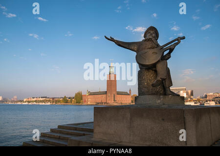 Eine Statue von Evert Taube Wiedergabe der Laute an der Waterfront auf Riddarholmen Insel, mit Rathaus Gebäude im Hintergrund, Stockholm, Schweden. Stockfoto