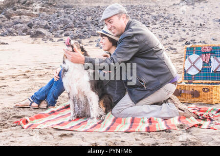 Paare, die in Liebe und Partnerschaft am Strand bei einem Picknick mit bester Freund der Hund mit Ihnen in spielerische Tätigkeit. Glück und tiergestützte Therapie lif zu genießen Stockfoto