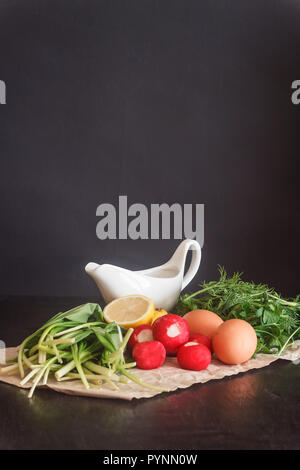 Bereit, frisches Gemüse und Produkte für Salat, Bärlauch, Radieschen, Zitrone, urkop, huhn eier, Sauce in einen Topf auf Pergamentpapier auf einem dunklen Ba Stockfoto