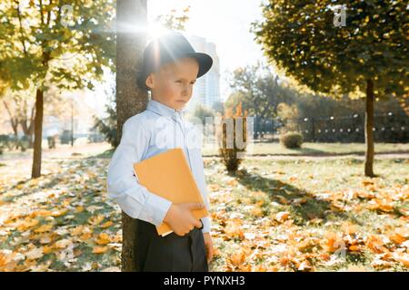 Beleidigt little boy in hat, klassische Shirt mit Buch in der Hand. Steht in der Nähe ein Baum im sonnigen Herbst Park, die goldene Stunde. Stockfoto