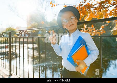 Wenig intelligente junge in Gläser mit Büchern, zeigt Zeigefinger nach oben Idee, Eureka. Hintergrund Herbst sonnigen Park, goldenen Stunde. Stockfoto