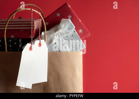 Zwei weiße ungeschriebene Labels hängen von einer Papiertüte voller bunter Geschenkboxen, auf einem roten Hintergrund. Stockfoto