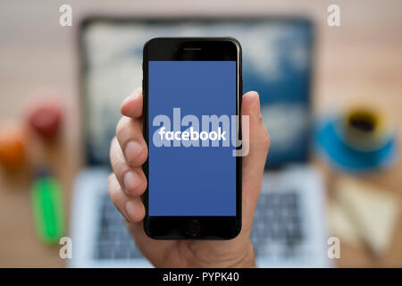 Ein Mann schaut auf seinem iPhone. das Facebook Logo, während saß an seinem Computer Schreibtisch (nur redaktionelle Nutzung). Stockfoto