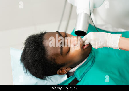 Frau Zahnarzt mit x-ray Maschine, Patienten, der auf Stuhl in der Zahnheilkunde. Junge afrikanische amerikanische Mann mit schlechten Zähnen. Medizin, Gesundheit, Stomatologie Konzept. Stockfoto