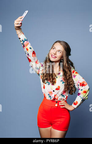 Studio Portrait von schönen trendy Mädchen mit langen lockigen Haar und Make-up Haltearm auf Taille und unter selfie per Handy über violette Hintergrund Stockfoto