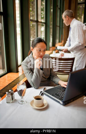 Geschäftsfrau auf ihrem Laptop in einem Cafe mit einem männlichen Kellner Einstellung dem Tisch hinter ihr zu arbeiten. Stockfoto