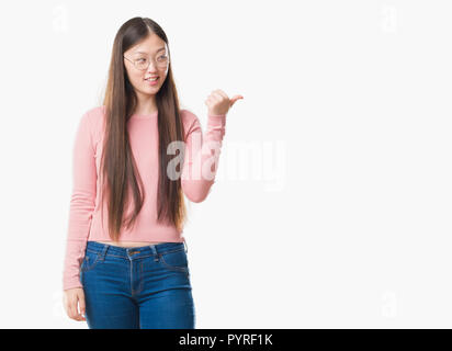Junge chinesische Frau über isolierte Hintergrund Brille lächelnd mit glücklichen Gesicht und zeigt auf die Seite mit dem Daumen nach oben. Stockfoto