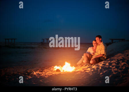 Glückliches junges Paar kuscheln zusammen neben einem romantisch leuchtenden Lagerfeuer am Strand bei Nacht. Stockfoto