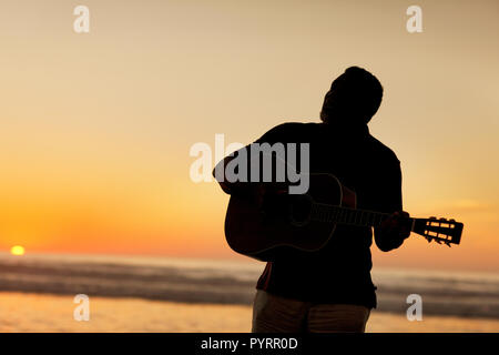 Gerne älterer Mann spielt eine akustische Gitarre am Strand bei Sonnenuntergang. Stockfoto