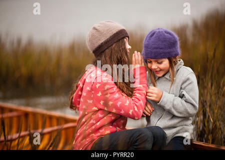 Zwei Schwestern weiterempfehlen Geheimnisse und lachen, als sie zusammen in einem hölzernen Kanu zwischen Schilf Schilf am Ufer sitzen. Stockfoto