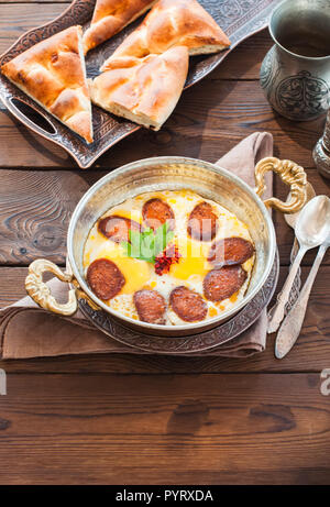 Türkisches Frühstück - Spiegeleier mit Würstchen (sucuk) und Gewürzen in einer Pfanne auf einem hölzernen Hintergrund. Stockfoto